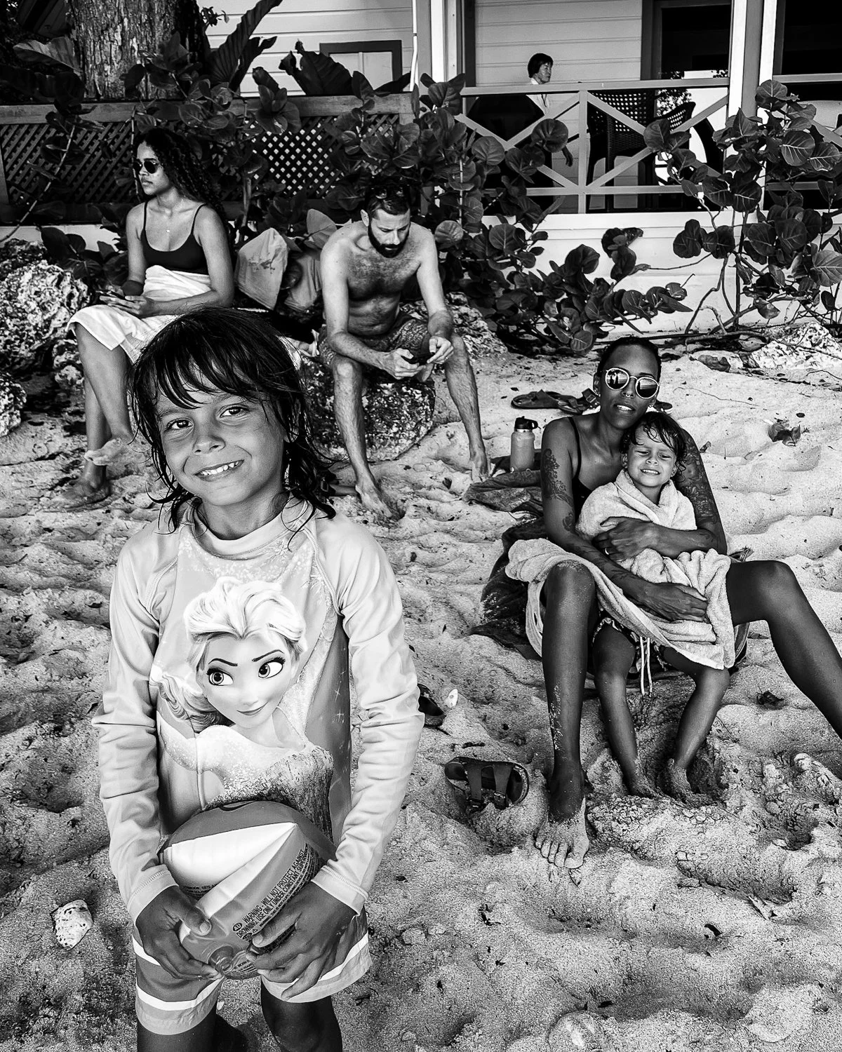 A group of people relaxing on a beach, including a smiling young girl holding a volleyball in the foreground, a woman and a young girl sitting on the sand, a shirtless man sitting on rocks looking at his phone, and a woman sitting behind them. There