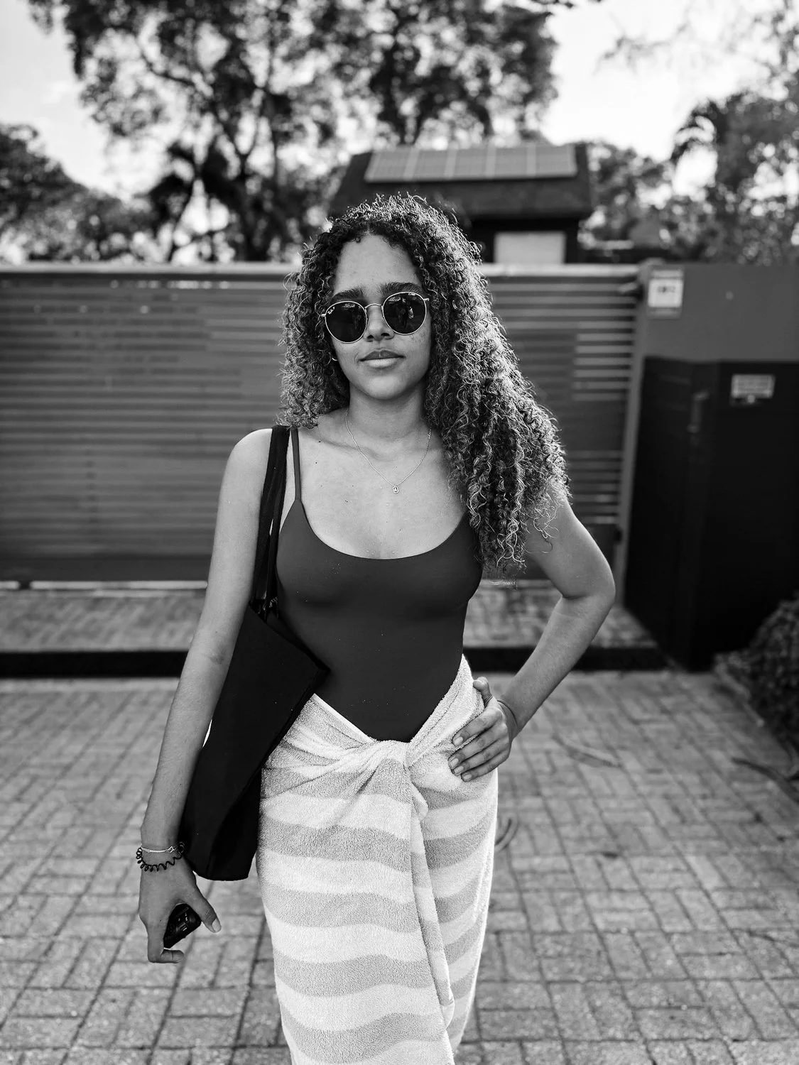 A woman with curly hair wearing sunglasses, a sleeveless top, and striped pants, standing outdoors in front of a fence with trees and a house in the background.