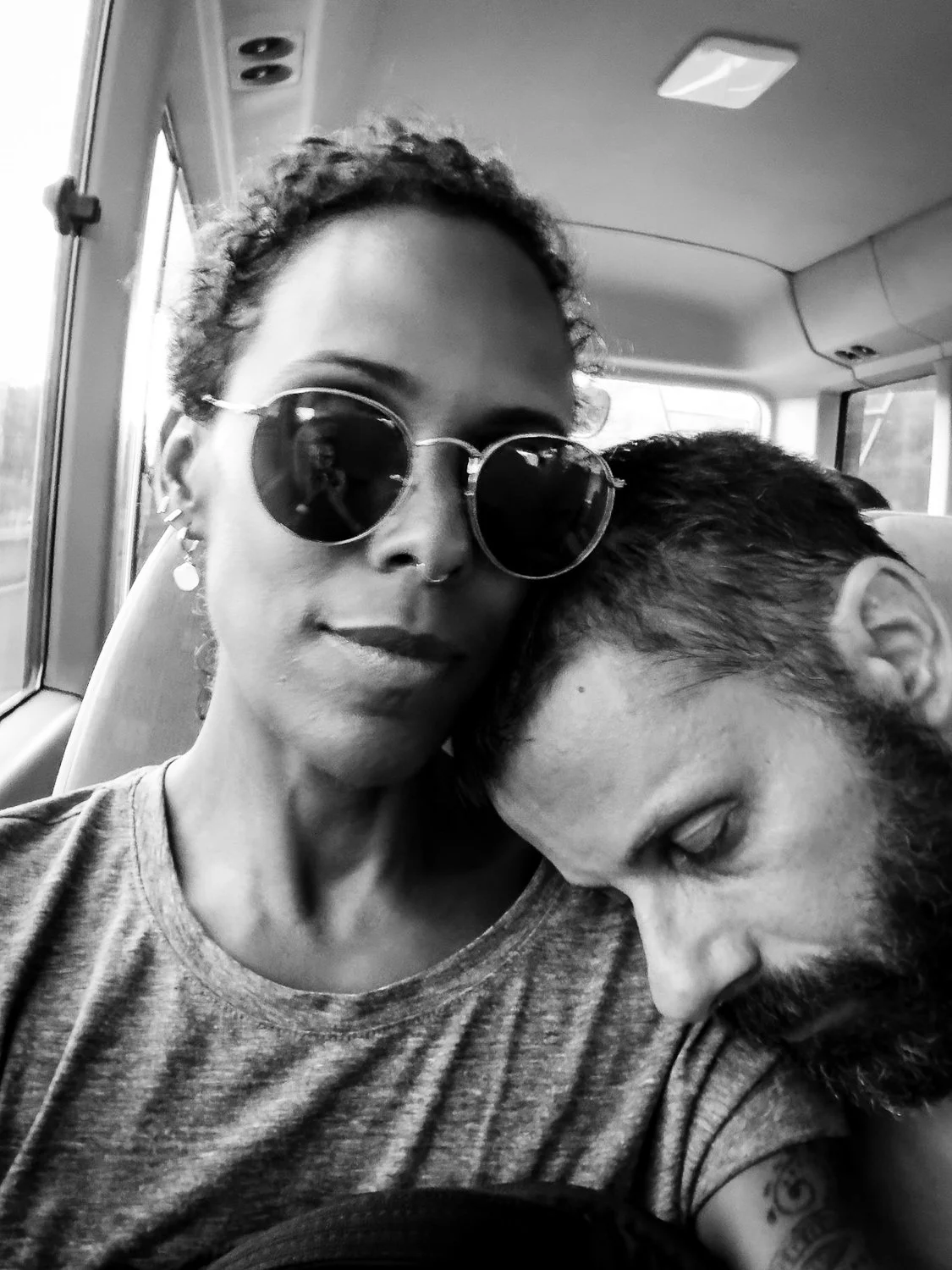 A black-and-white photo of a woman with sunglasses and short hair sitting next to a man with a beard and tattoos, resting his head on her shoulder on a bus.