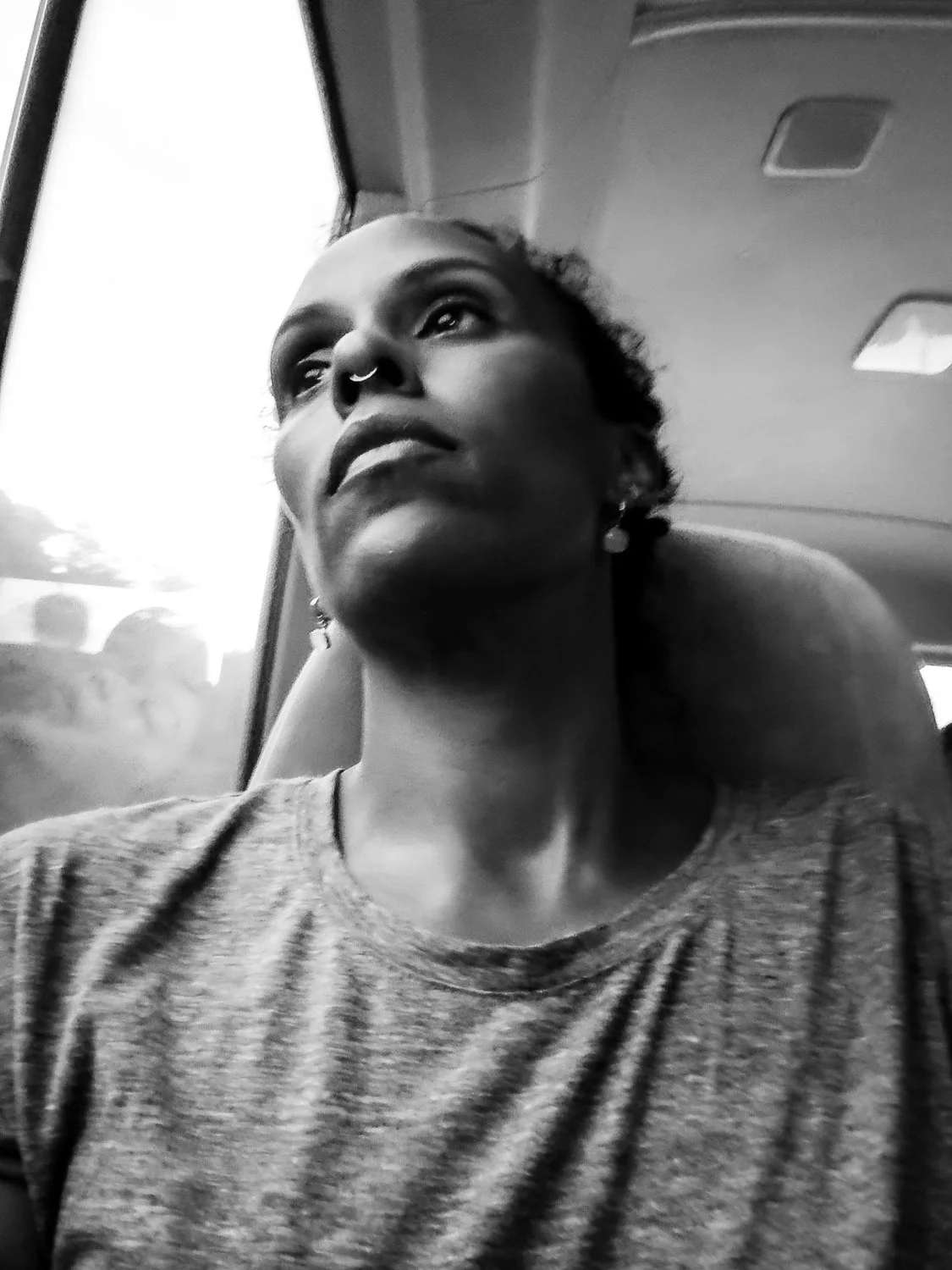 A woman with short hair, wearing earrings and a t-shirt, sitting on a bus or train, looking out the window with a thoughtful expression.