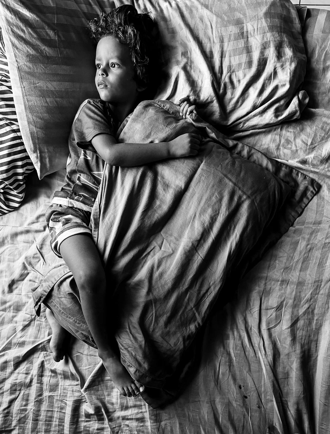 A young child lying in bed, covered with a blanket, looking off to the side with a thoughtful expression.