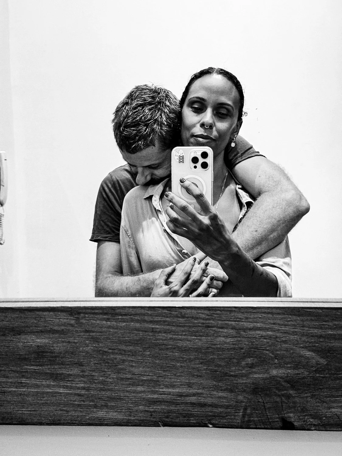A black and white photo of a couple taking a mirror selfie, with the man hugging the woman from behind, both appearing affectionate.
