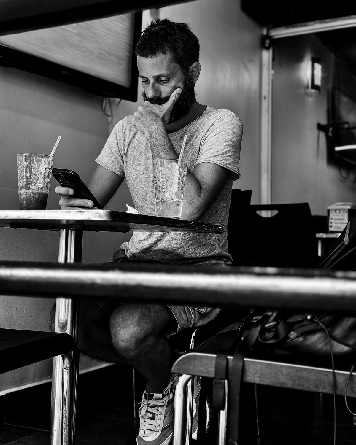 A man with a beard sitting at a table, looking at his cellphone, with two drinks and straws in front of him, in a cafe or restaurant setting.