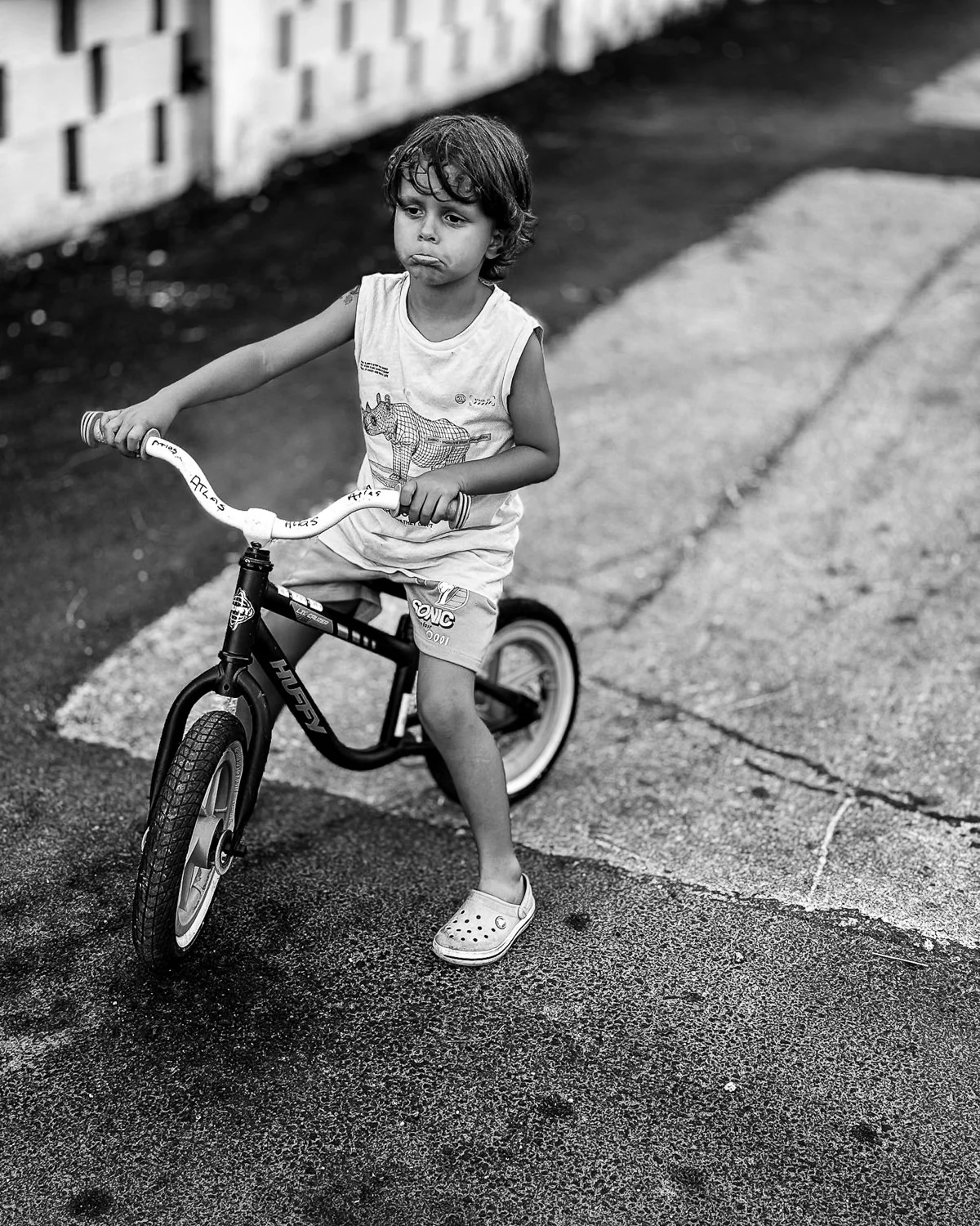 A young boy with curly hair riding a small bicycle on a paved path, looking ahead with a serious expression, wearing a sleeveless shirt, shorts, and Crocs.