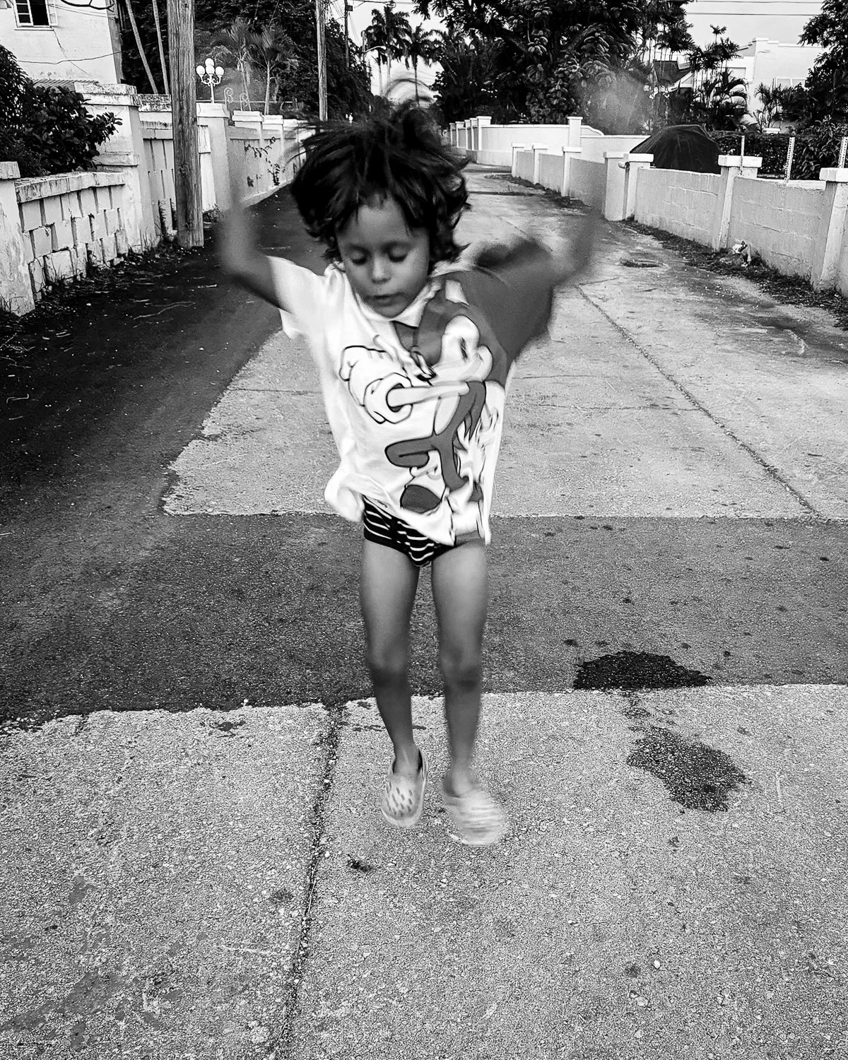 Child jumping on a paved street, wearing a cartoon character t-shirt, striped shorts, and sunglasses, with a white fence and trees in the background.