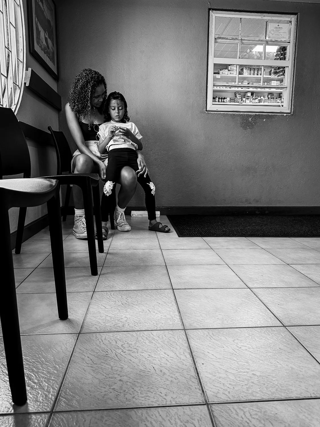 A woman and a girl sitting on chairs in a waiting area, looking at a phone. The room has tiled flooring, chairs, and a window with a view of items behind glass.