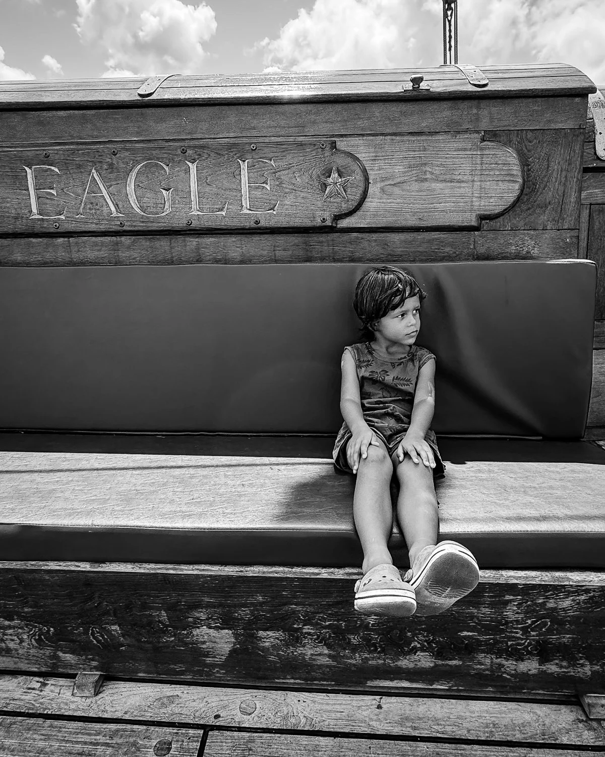 A young girl sitting on a wooden bench with her legs hanging over the edge, looking to her right with a pensive expression. Behind her, there is a large wooden structure with the word 'EAGLE' and a star carved into it, against a backdrop of clouds.