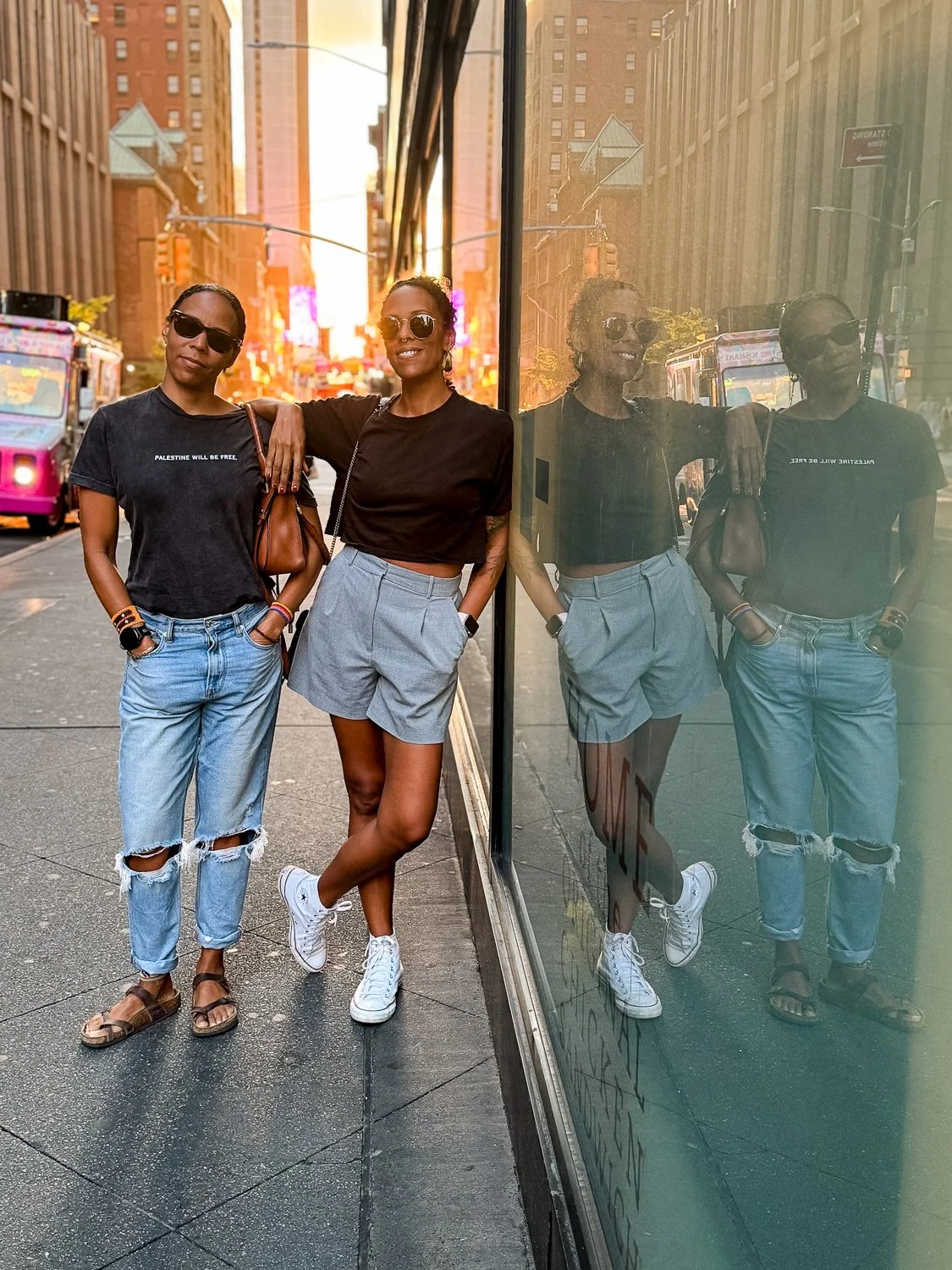 Three women stand on a city sidewalk next to a glass wall that reflects their image. They are dressed casually with sunglasses, and city buildings and a double-decker bus are visible in the background during sunset.