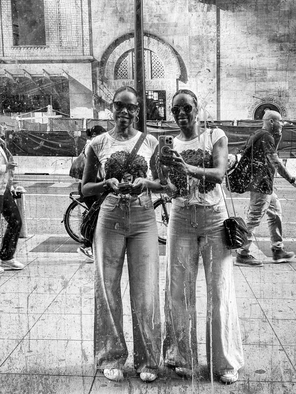 Two women in sunglasses taking a photo of themselves reflected in a rain-streaked glass window on a city street.