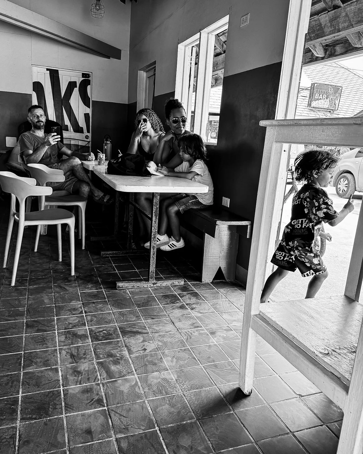 People sitting at a table inside a restaurant or cafe, with children playing near the entrance, black and white.