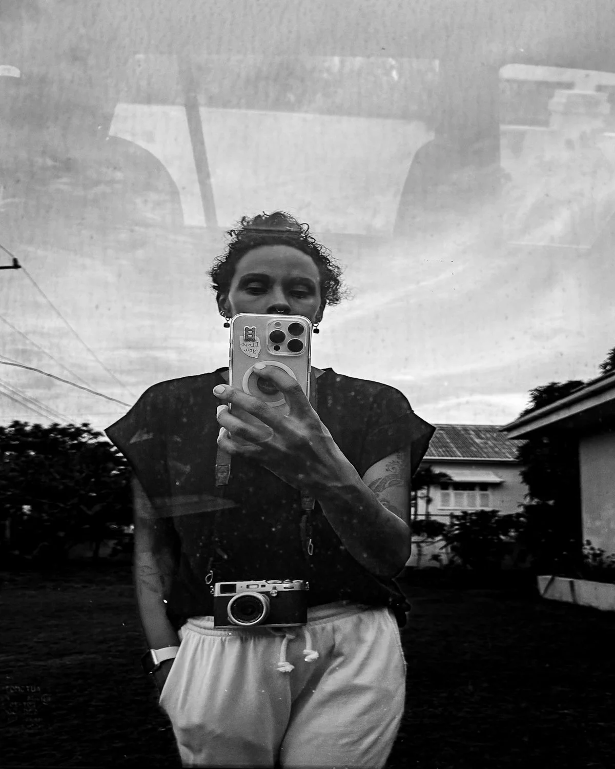 A person taking a selfie in a mirror outdoors on a cloudy day, holding a phone with a round grip, wearing a dark top and light-colored pants, with a camera hanging around their neck.