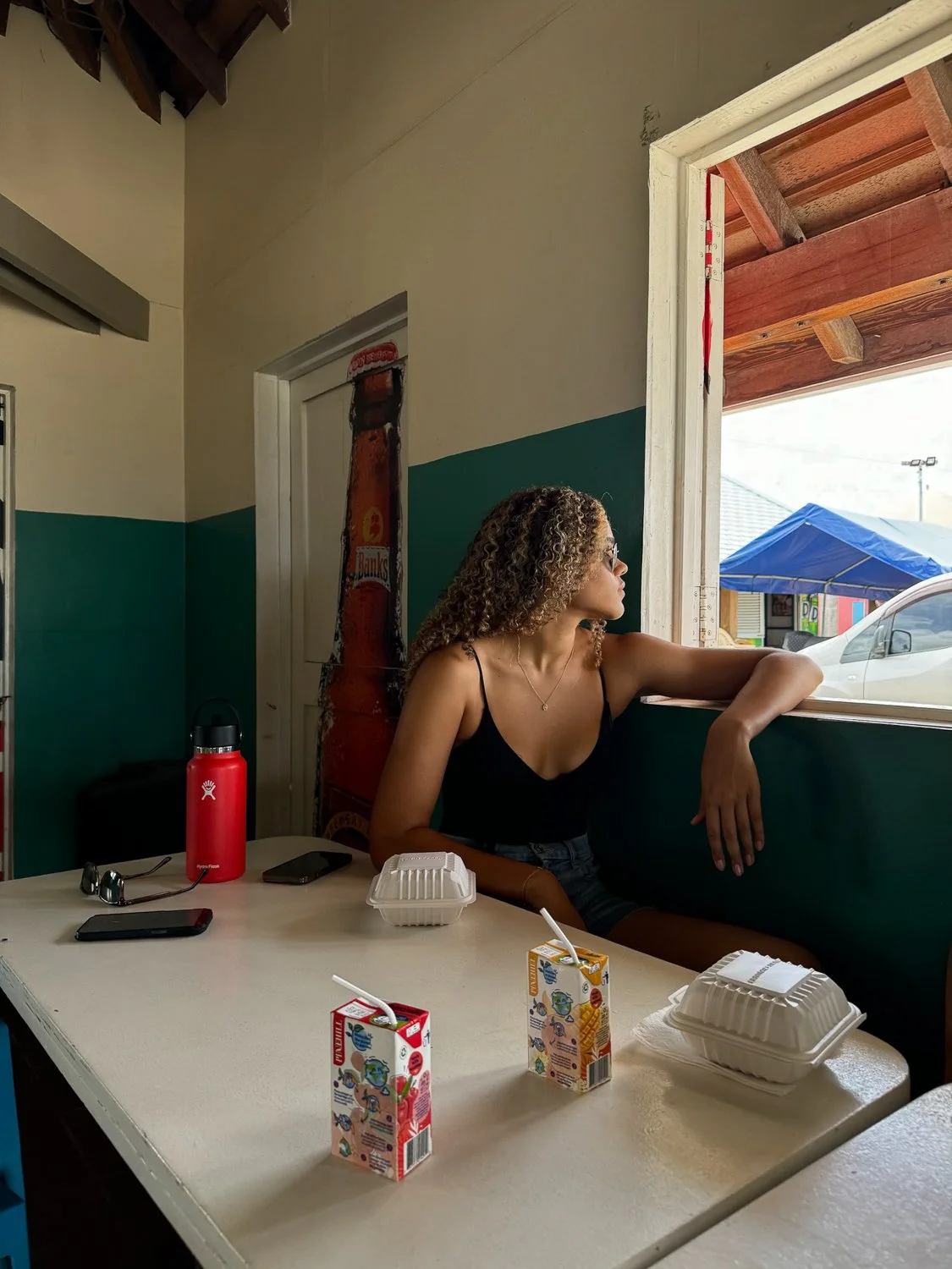 A woman sitting inside a restaurant by a window, looking outside. There are two cartons of juice, a foam takeout container, a red water bottle, and a cell phone on the table.