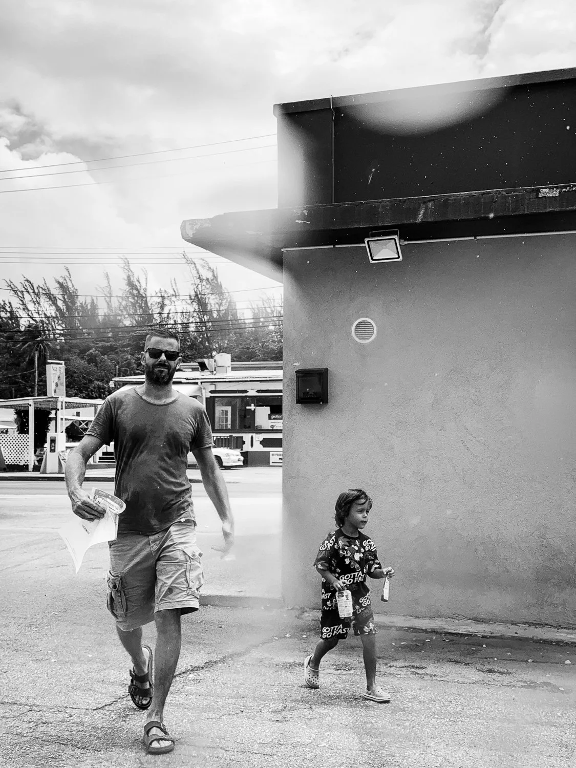 A man and a young girl walking outside near a building, with the man holding papers and the girl holding bottles, in a black and white photo.