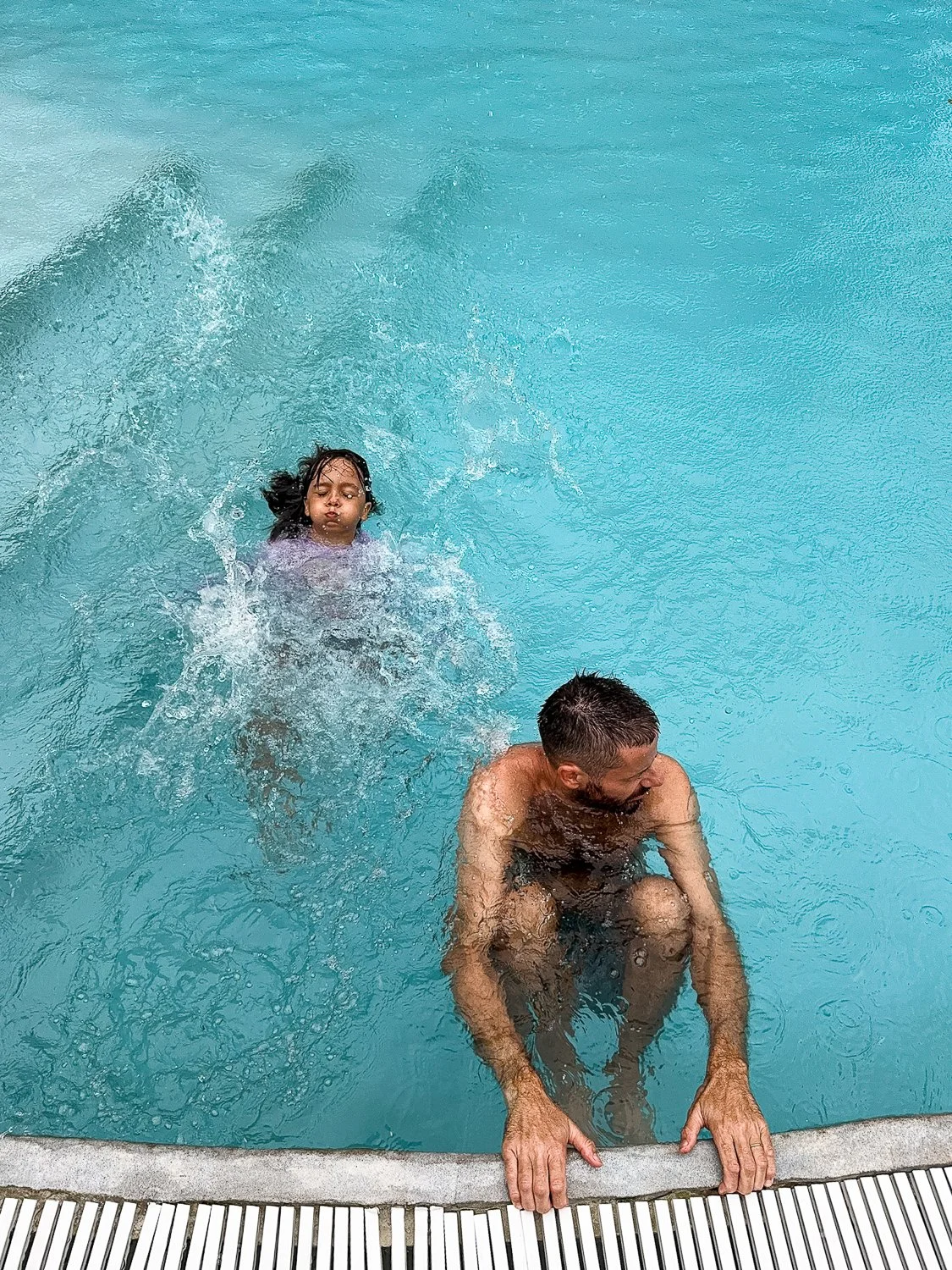A man and a young girl in a swimming pool, the girl is swimming and the man is crouching at the pool edge holding onto it.