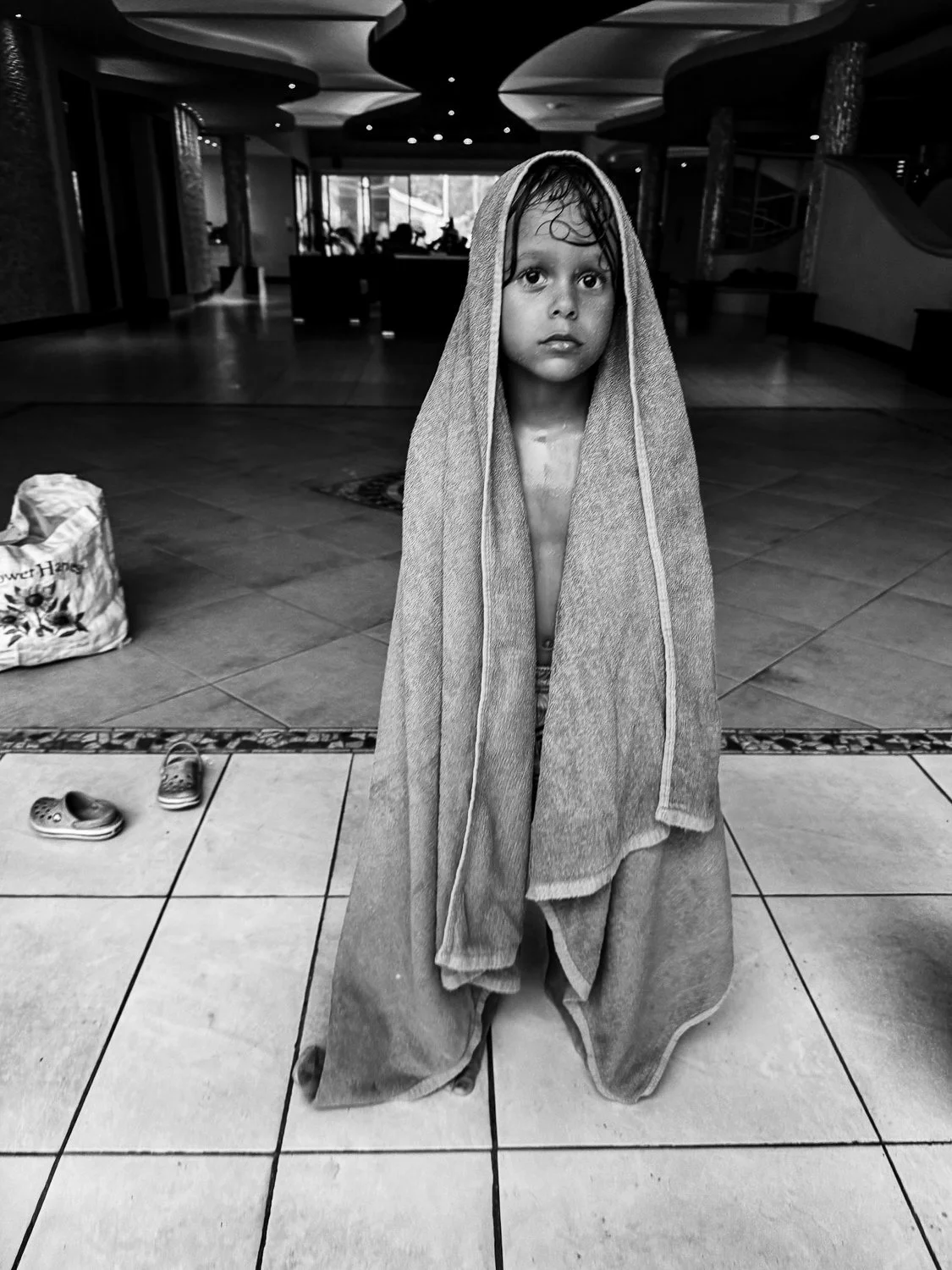 A young child with wet hair, standing inside a building, wrapped in a towel and looking at the camera.
