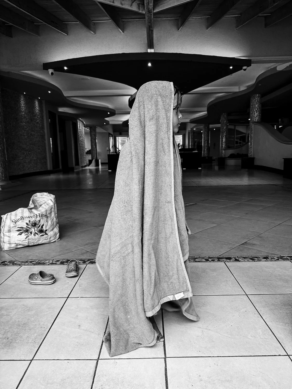 A person standing indoors with a towel draped over their head and shoulders, facing to the right. Shoes are on the floor nearby, and a bag labeled 'Sunflower Honey' is also on the floor. The background shows a spacious, well-lit interior with modern