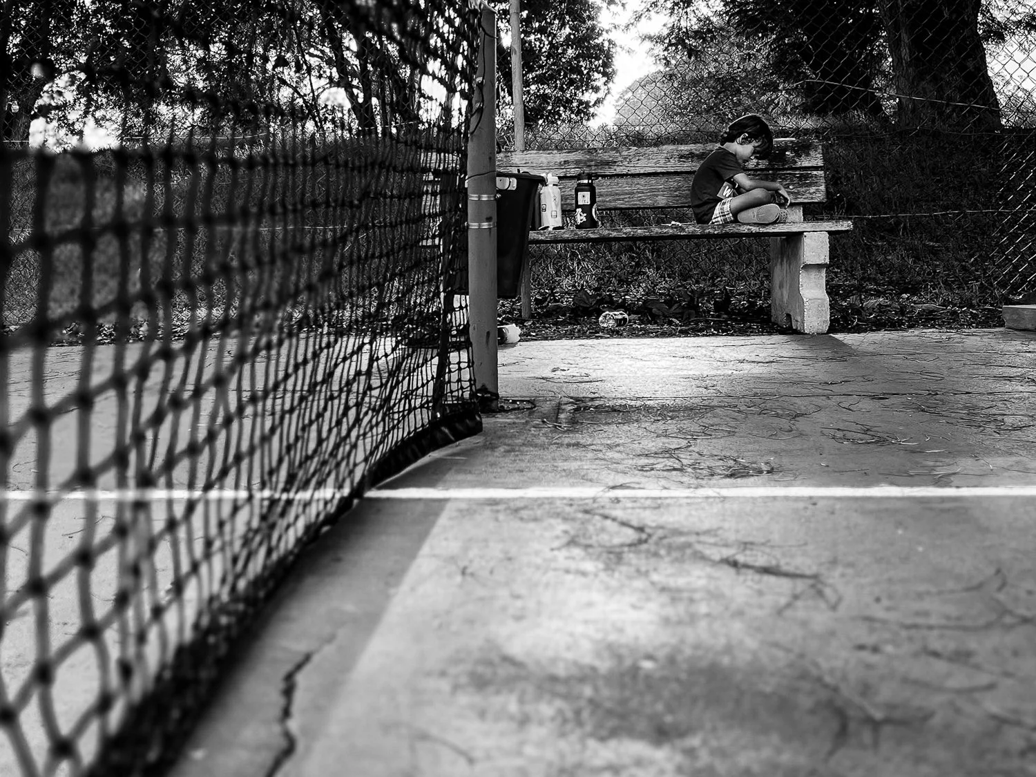 A black and white photo of a young boy sitting alone on a wooden bench, looking down with his legs crossed. The scene is outside a fenced tennis court, with a chain-link fence on the left side of the image, and some trees in the background.