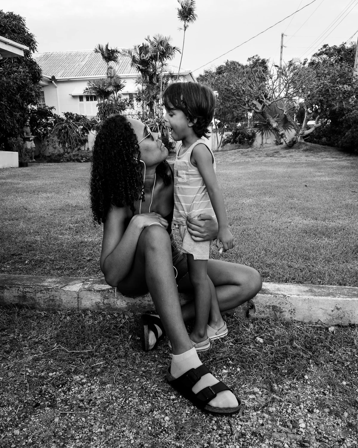 A woman with curly hair and sunglasses sitting on the curb, holding a young boy by the waist, who is standing and facing her with his tongue out. They are outdoors in a yard with trees and a house in the background.