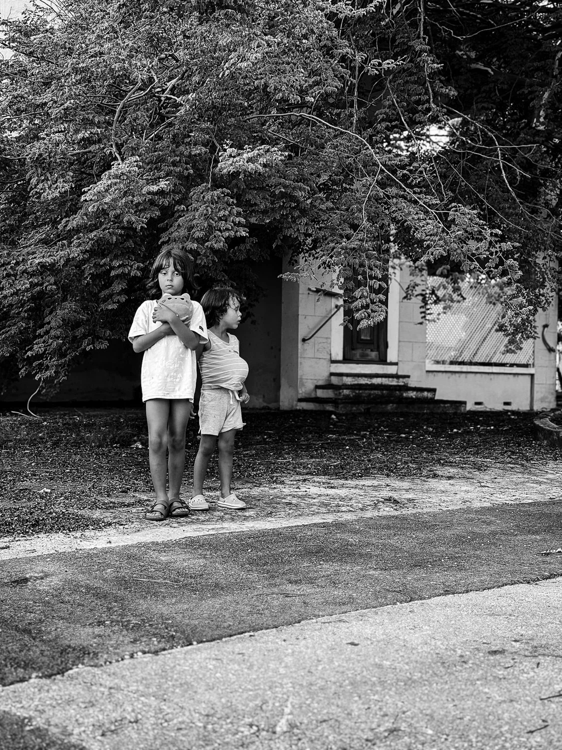 Two children, a girl holding a toy and a boy standing beside her, standing outdoors near a house with trees overhead, captured in black and white.