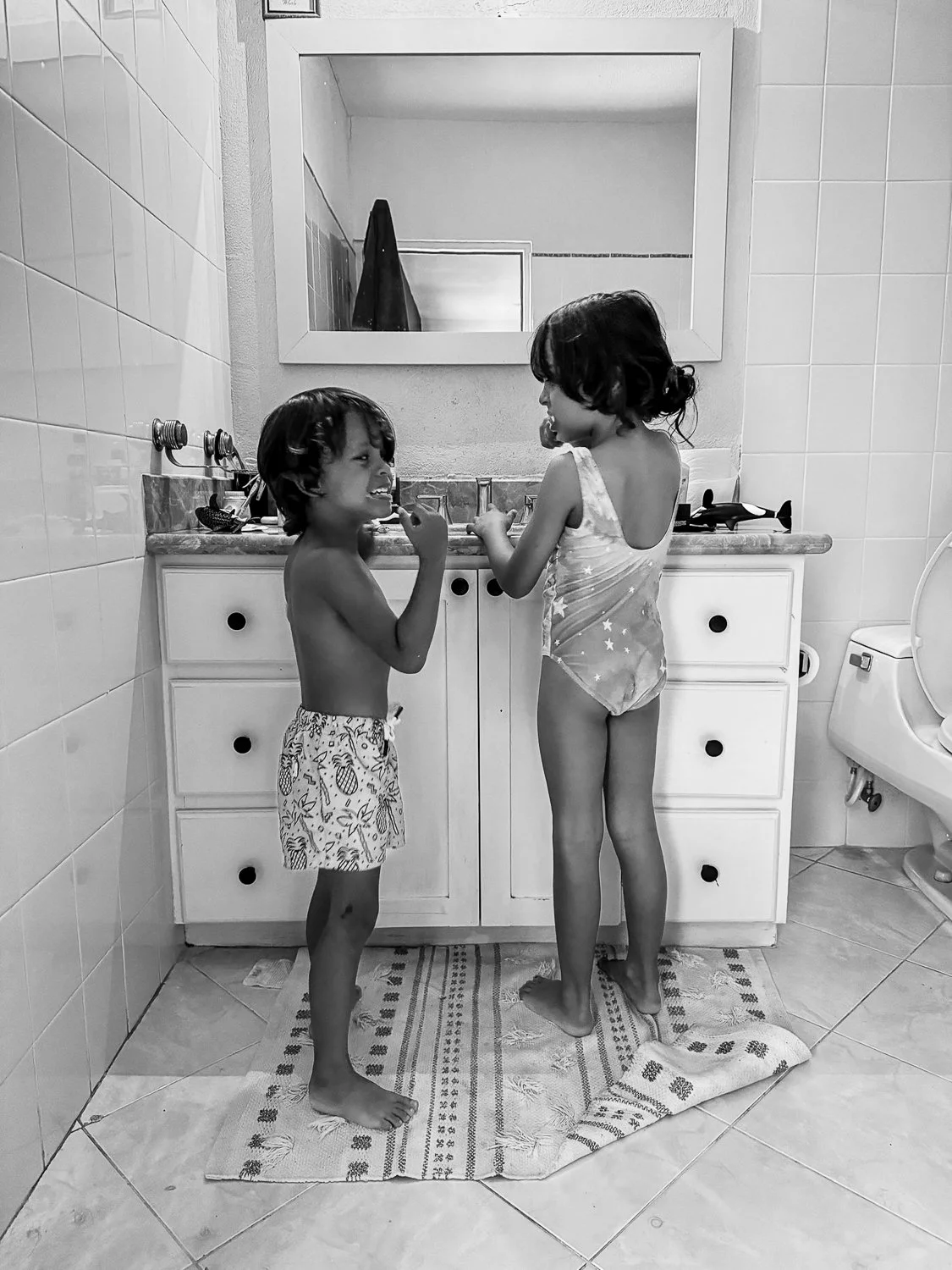 Two young children, a boy and a girl, are standing on a bathroom rug in front of a bathroom vanity mirror. The boy is shirtless and wearing patterned shorts, smiling and looking at the girl. The girl is wearing a star-patterned nightgown and facing t
