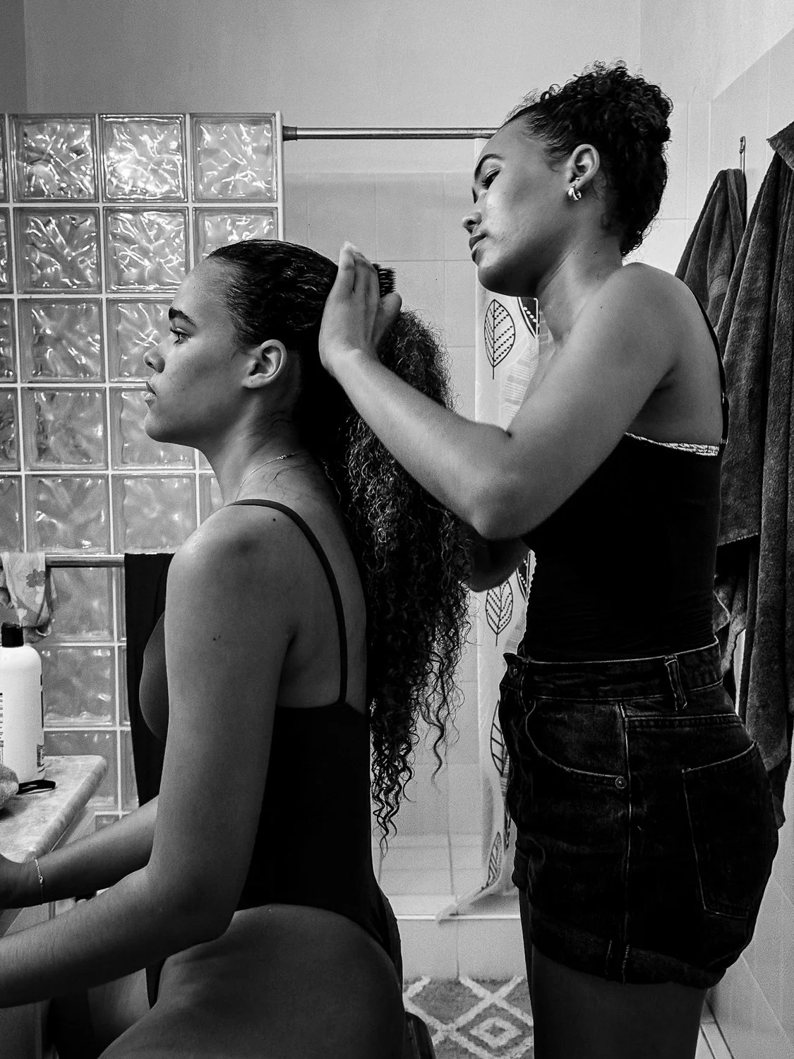 Two women in a bathroom, one sitting and the other styling her long curly hair.