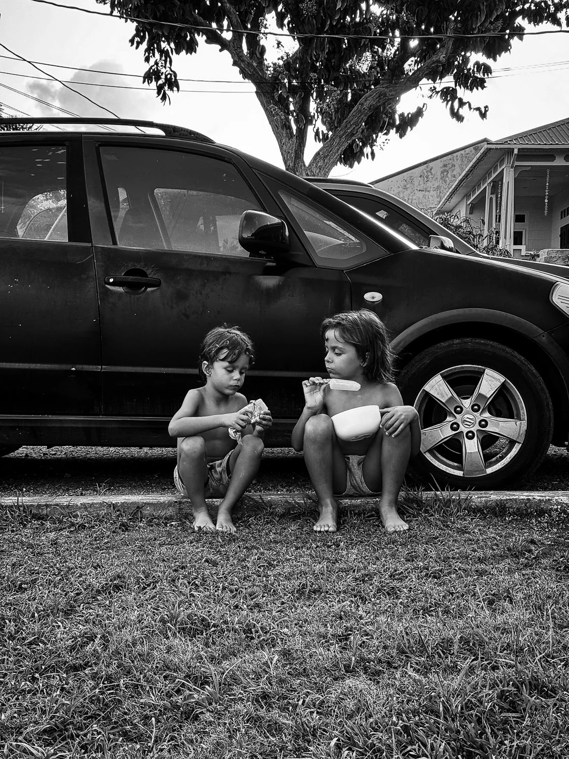 Two children sitting on the curb next to a parked car, eating snacks. One girl is holding a popsicle, and a boy is holding food wrapped in foil. The scene is outdoor with grass in the foreground and trees and houses in the background, in black and wh