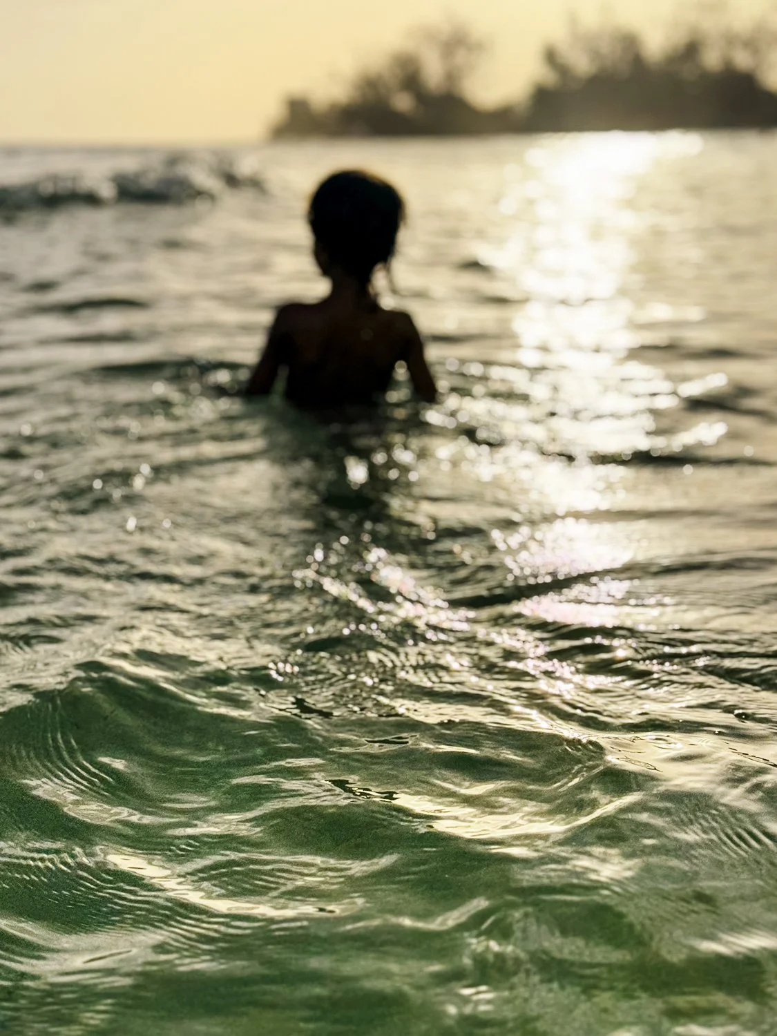 A child standing in the ocean during sunset, silhouette with the sun reflecting on the water.