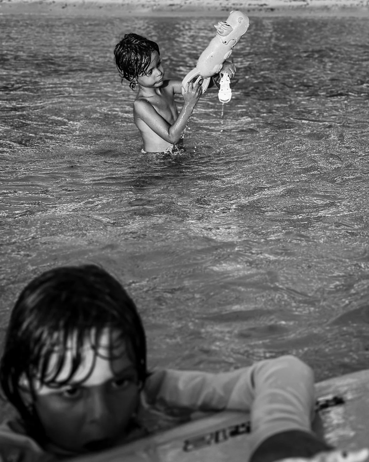 A black-and-white photograph of two children playing in the water at a beach, one child is in the water holding a toy gun, and another is resting on a floating object, partially submerged, with wet hair and an expression of focus.