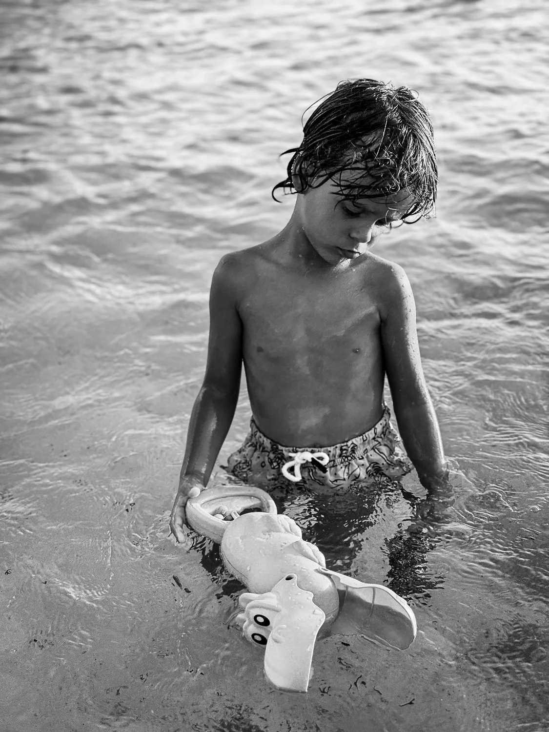 A young boy with wet hair standing in the shallow water at the beach, holding a toy octopus in his hand, looking down.