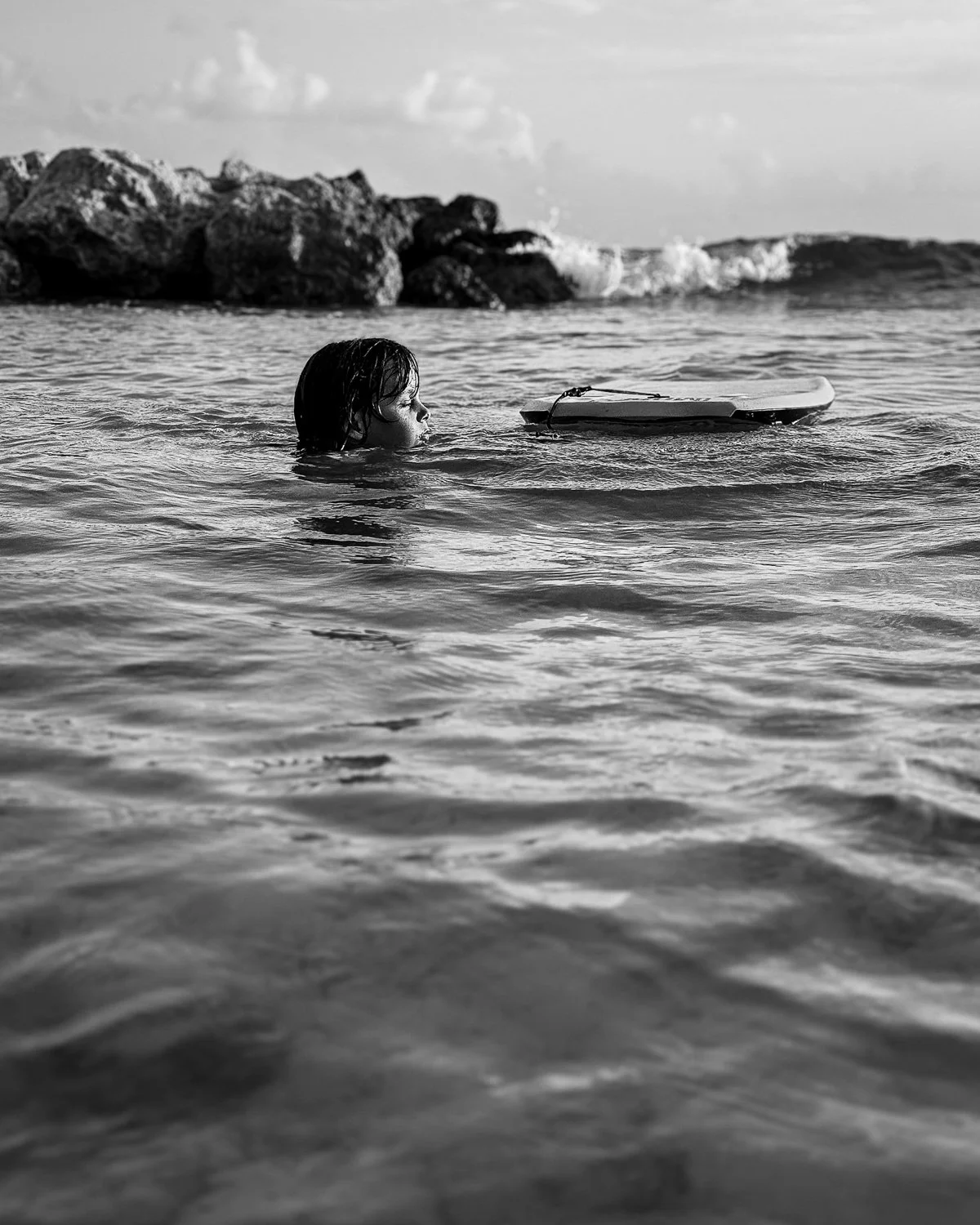 A child with wet hair in the water looking at a paddleboard near a rocky shoreline, black and white photo.
