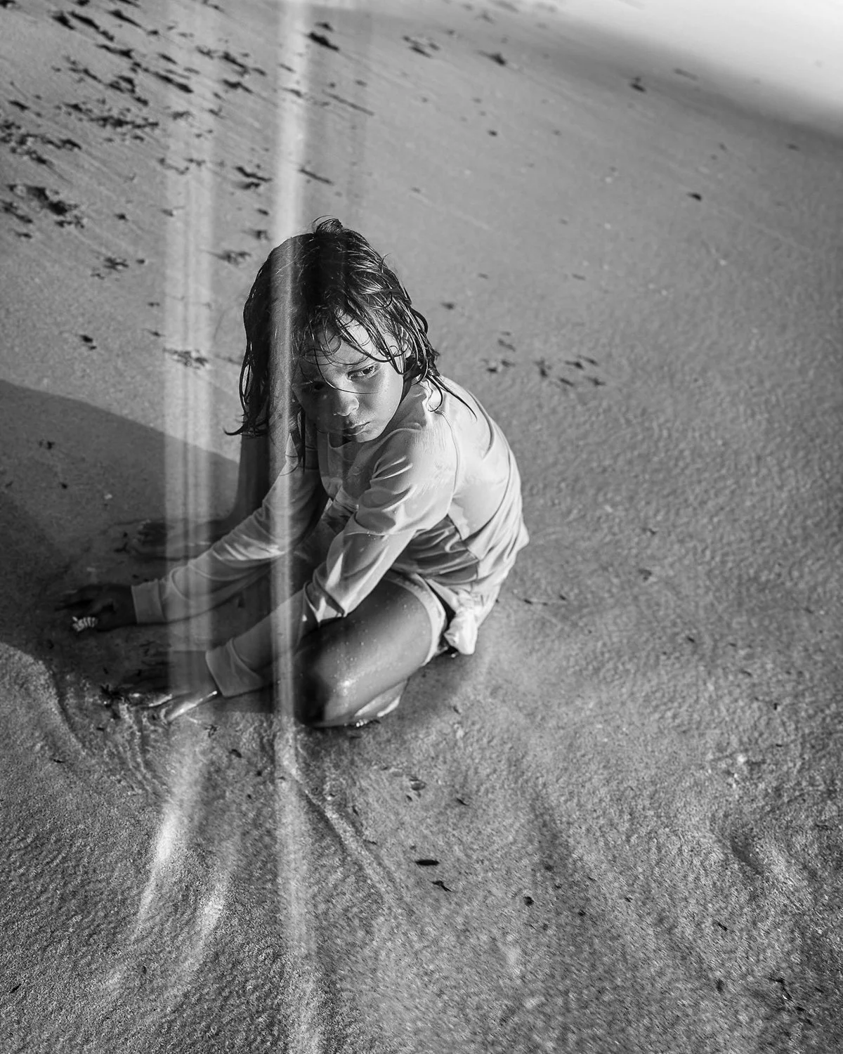 A young girl with wet hair sits on the wet sand at the beach, looking back with a serious expression, illuminated by sunlight and a vertical light streak.