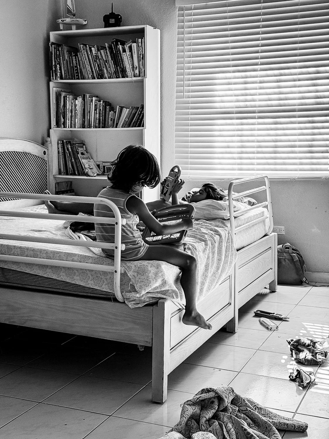 A black and white photo of two children on a bed in a bedroom. One child is sitting at the edge of the bed with legs dangling, holding a tablet and a video game controller. The other child is lying on the bed, also holding a tablet. The room has a bo