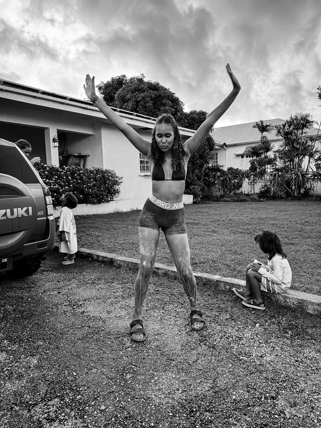 A woman standing outdoors with her arms raised, covered in paint or mud, wearing shorts and a sports bra, with children nearby in a yard outside a house, and a car parked to the left.