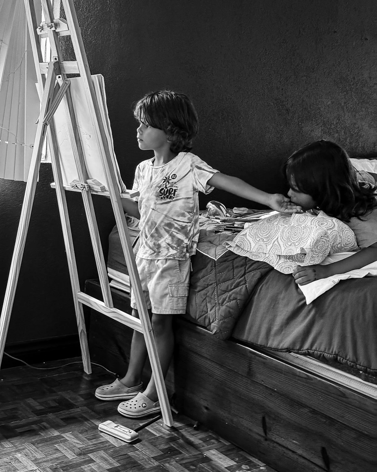 Two children are in a bedroom. One child is painting on an easel while the other child is lying on a bed, resting their head on a pillow.