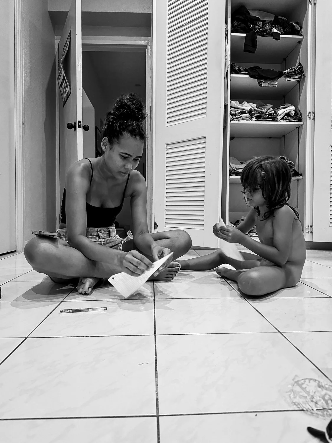 A black and white photo of a woman and a young girl sitting on a tiled floor, reading papers. They are inside a room with open closet doors filled with clothes, and a doorway behind them.