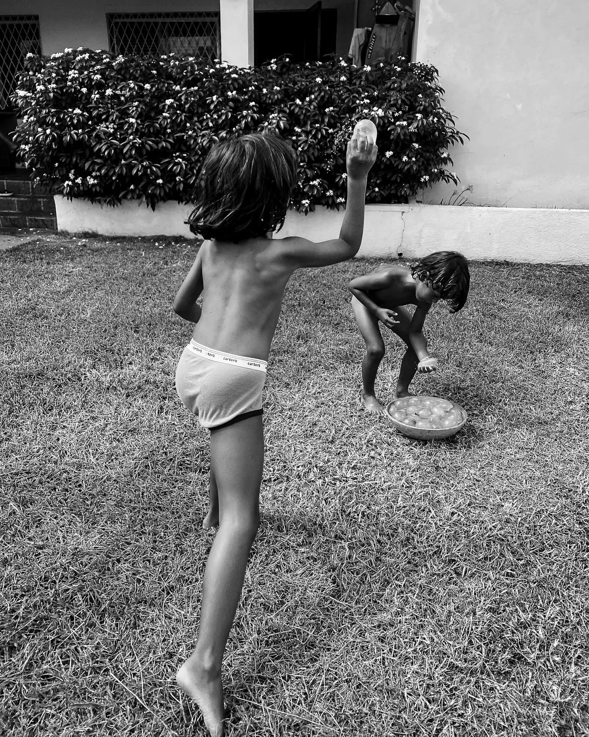 Two children playing on the grass, one standing and throwing a ball, the other crouching near a bowl of water balls, in an outdoor yard with bushes and a building in the background.