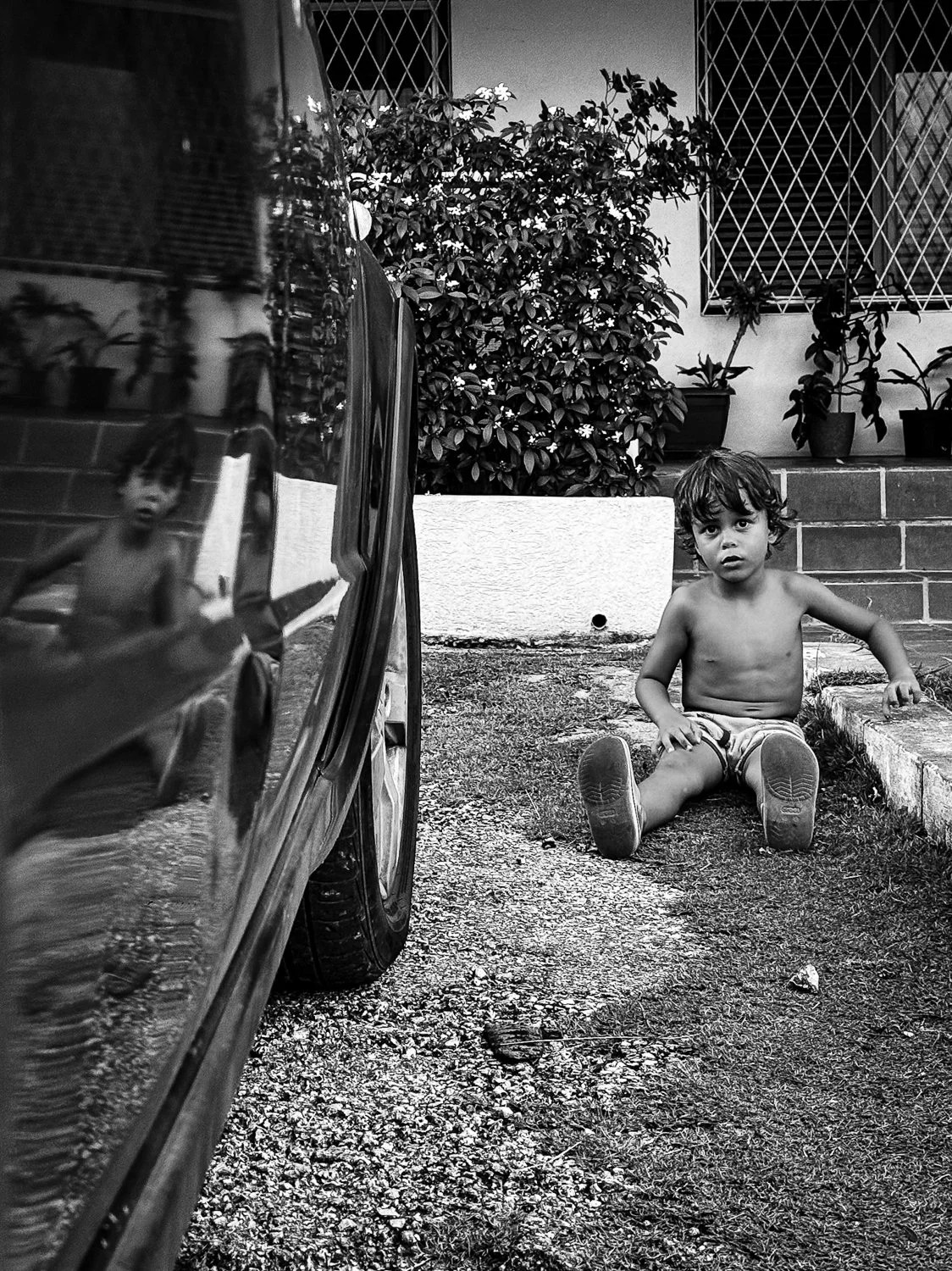 A young child sitting on the ground shirtless wearing shorts and shoes, looking at the camera, with a reflection of himself in the side of a parked car on the left. There are plants and a small staircase in the background.