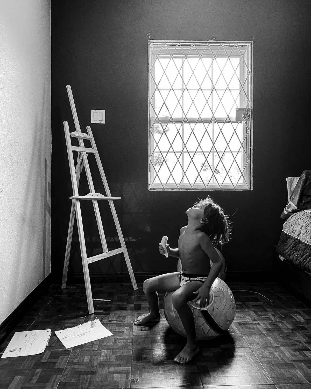 A young child sitting on an exercise ball in a room, looking up and smiling with arms raised, next to a bed and a window with security bars.