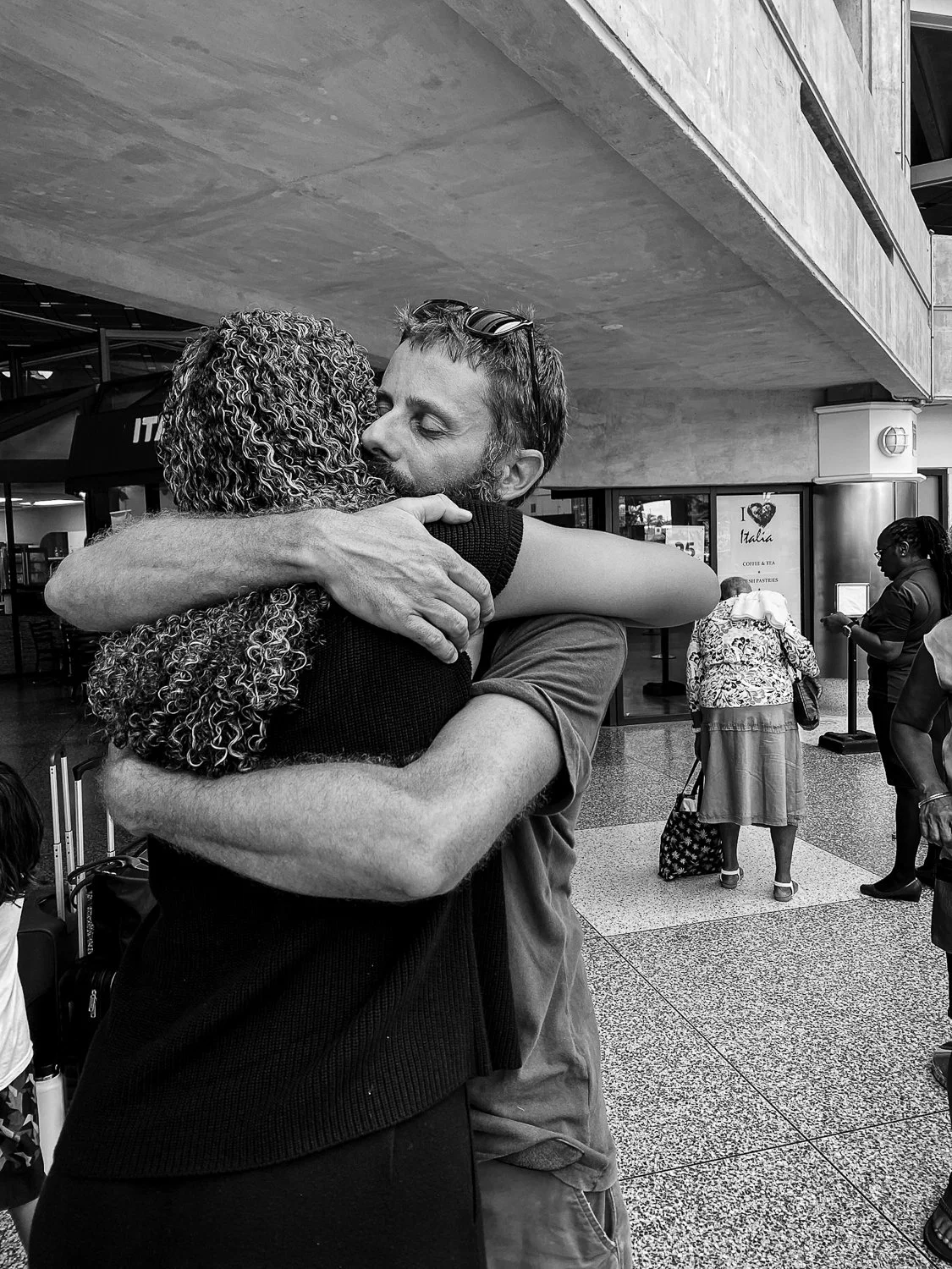 A man and woman hug at an airport or train station, with other travelers in the background.