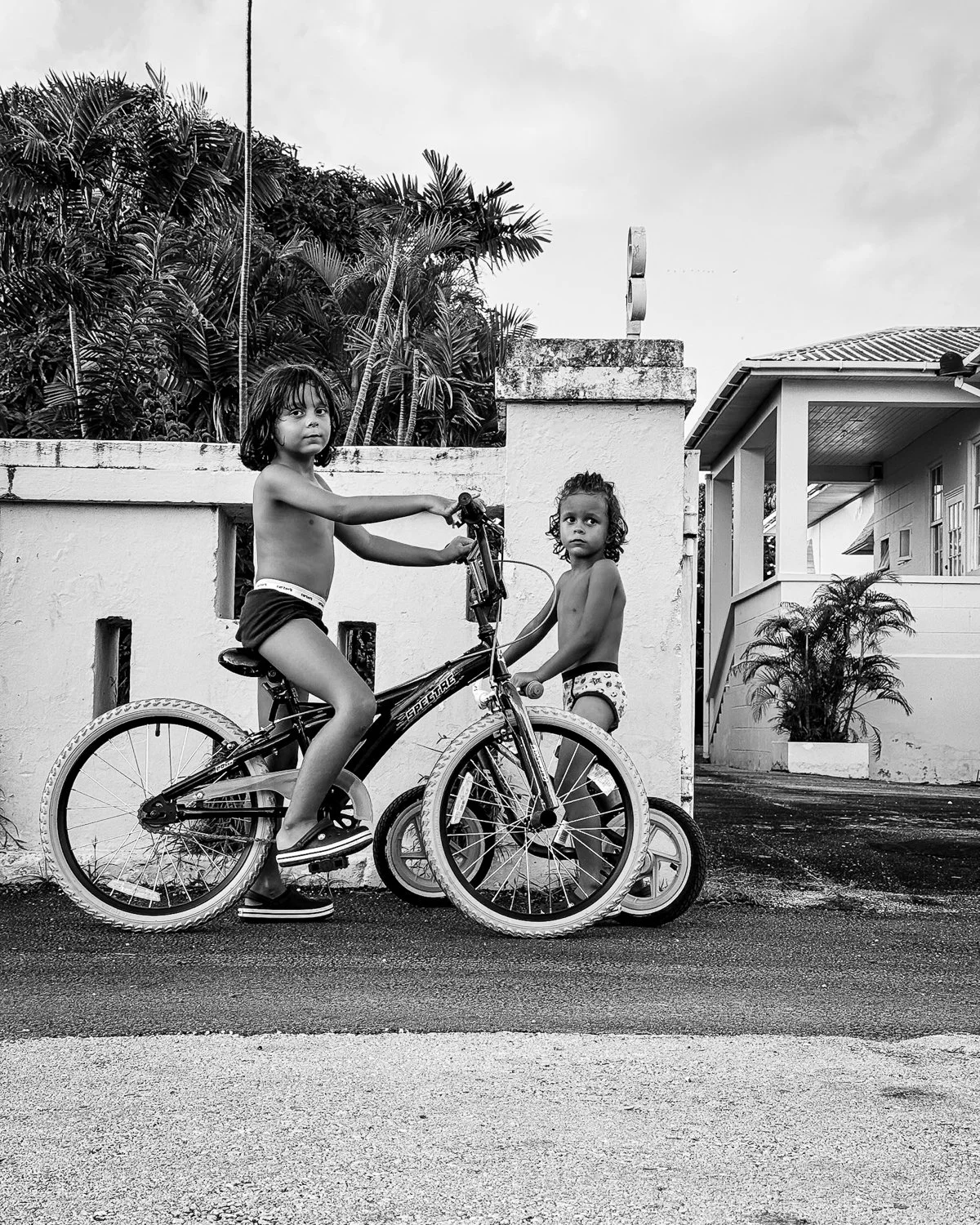 Two children without shirts and wearing underwear, one girl sitting on a bicycle and the other standing next to it, on a sidewalk in front of a white house with tropical plants in the background, black and white photo.