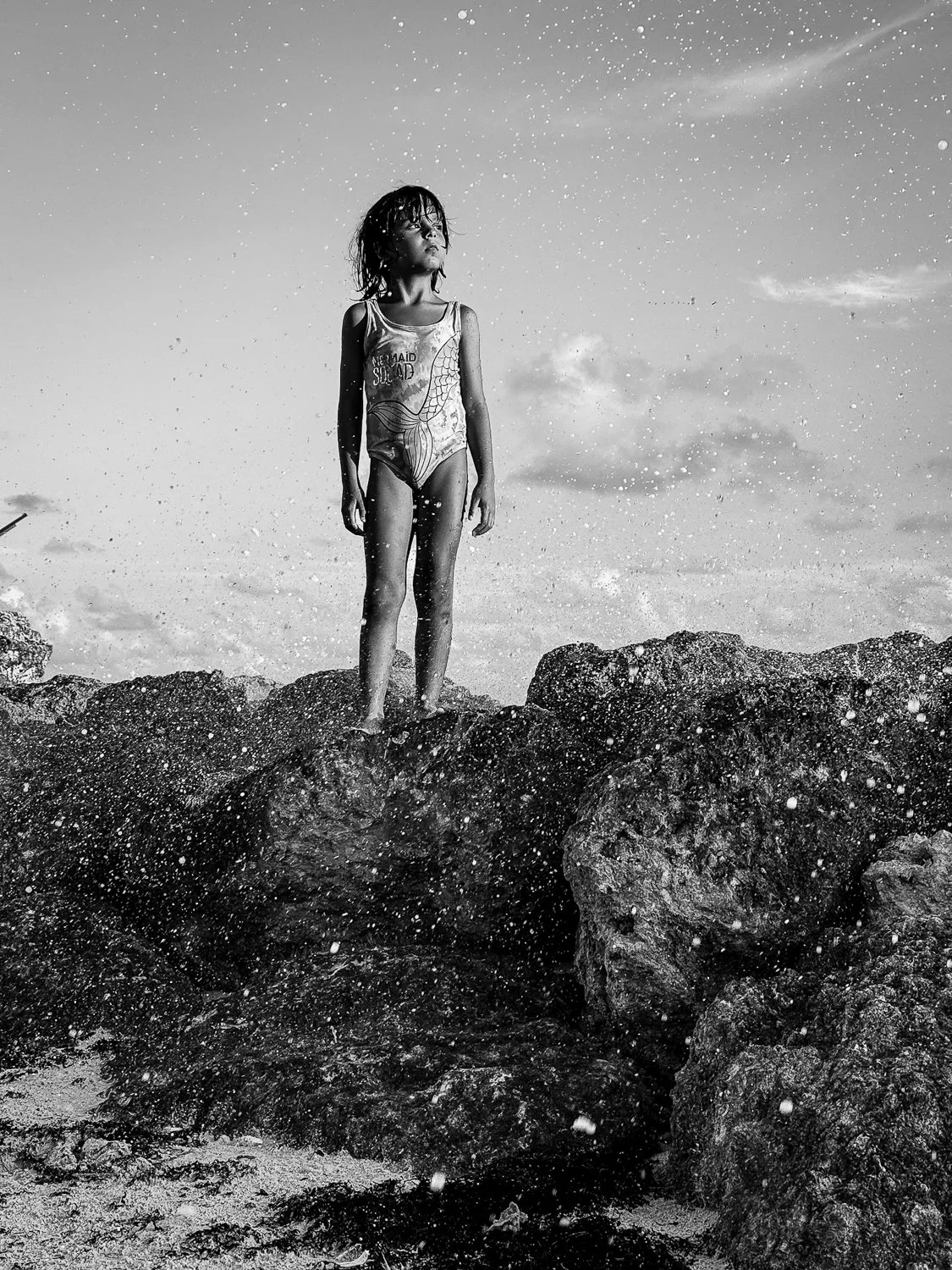 A young girl standing on rocks by the water, wearing a swimsuit, with water splashing around her under a cloudy sky.