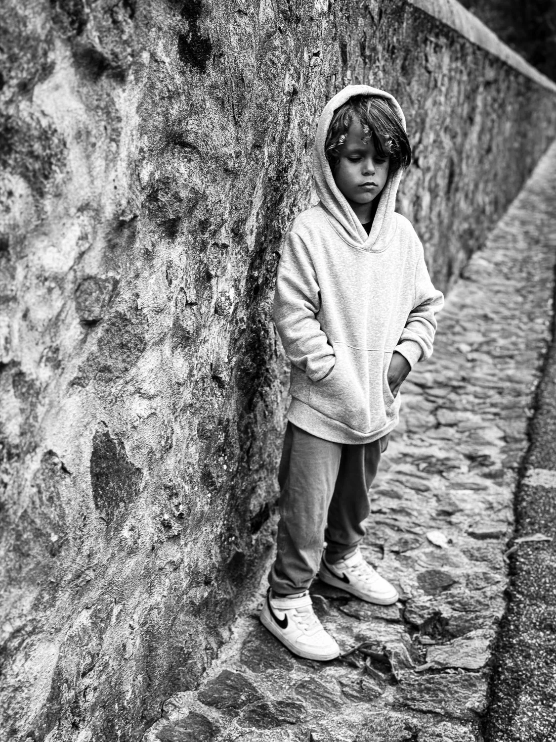 A young boy standing with his hands in the pockets of his hoodie, leaning against a stone wall on a cobblestone street, looking down with a serious expression.