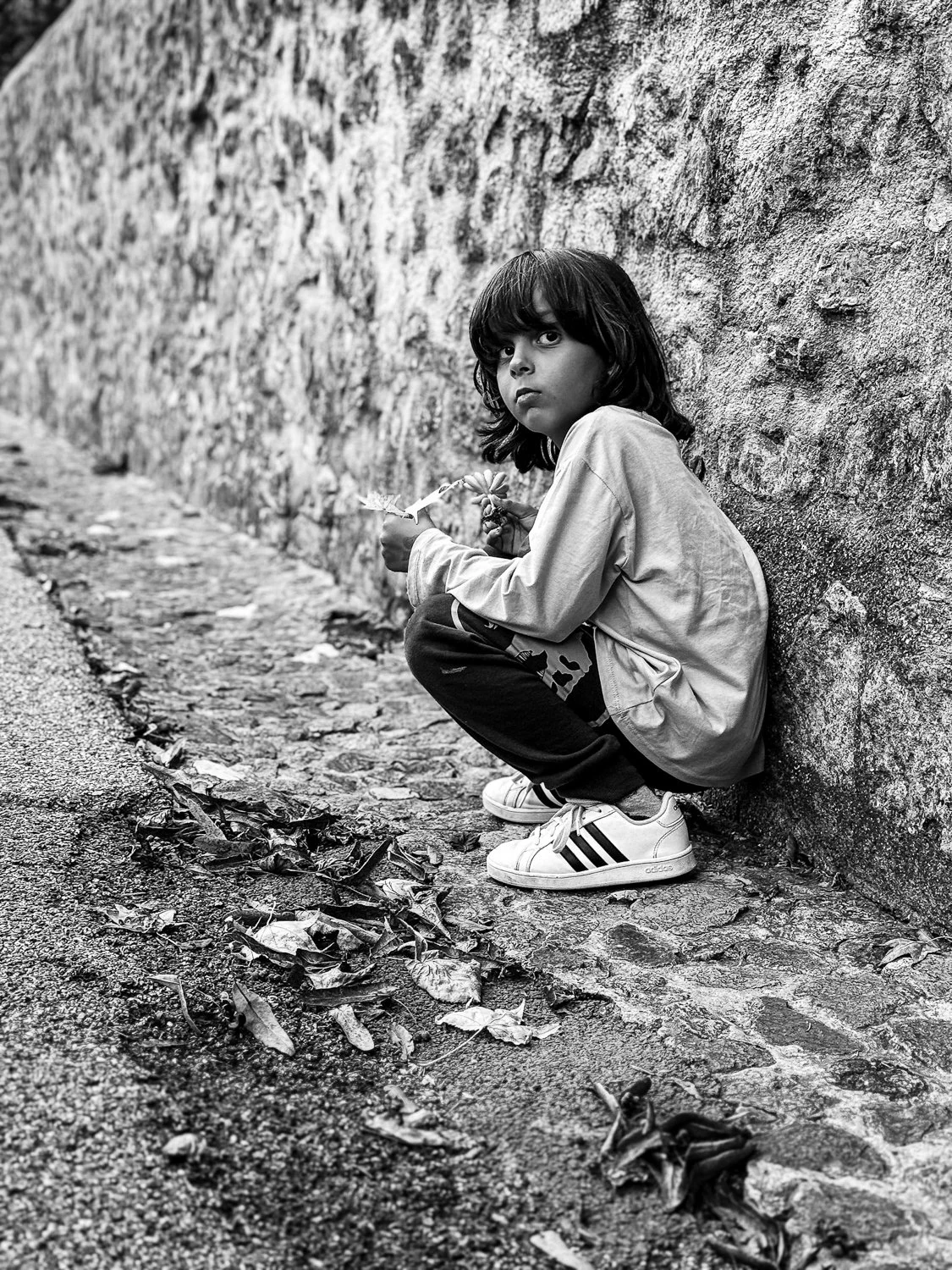 A young boy with dark hair squats on a cobblestone sidewalk, leaning against a textured stone wall, holding a toy or object, looking directly at the camera.