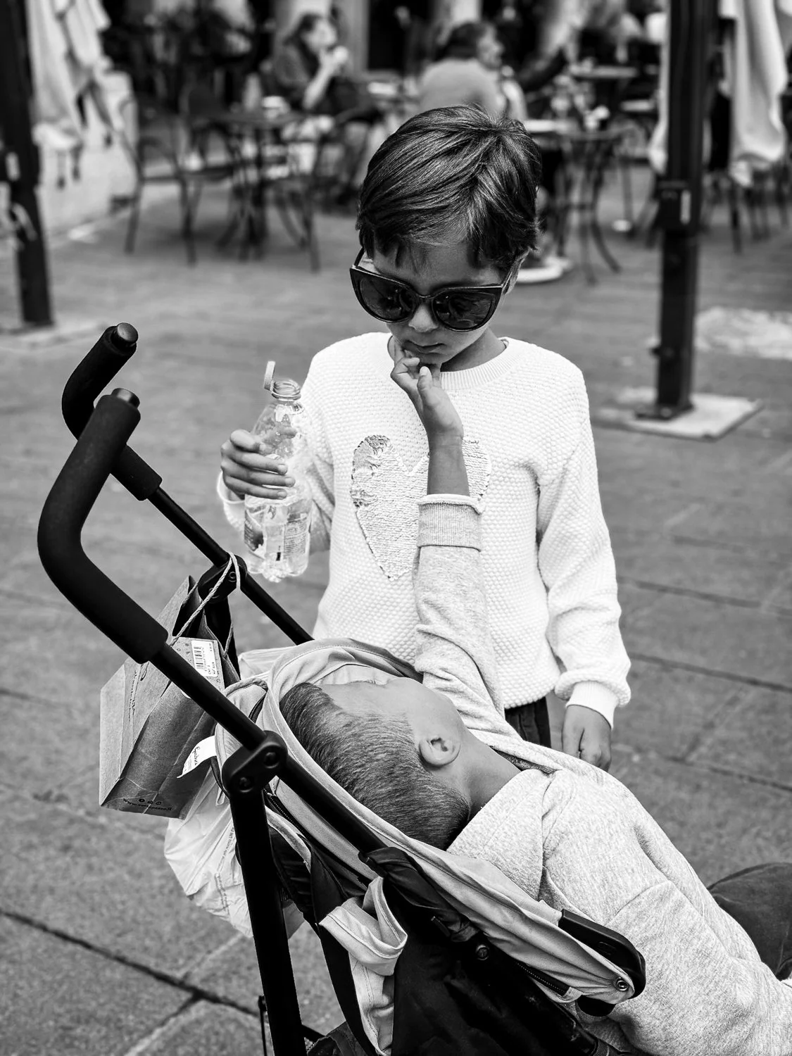 A young girl wearing sunglasses and a white sweater stands next to a stroller with an unconscious child, touching the child's face gently, in a busy outdoor setting.