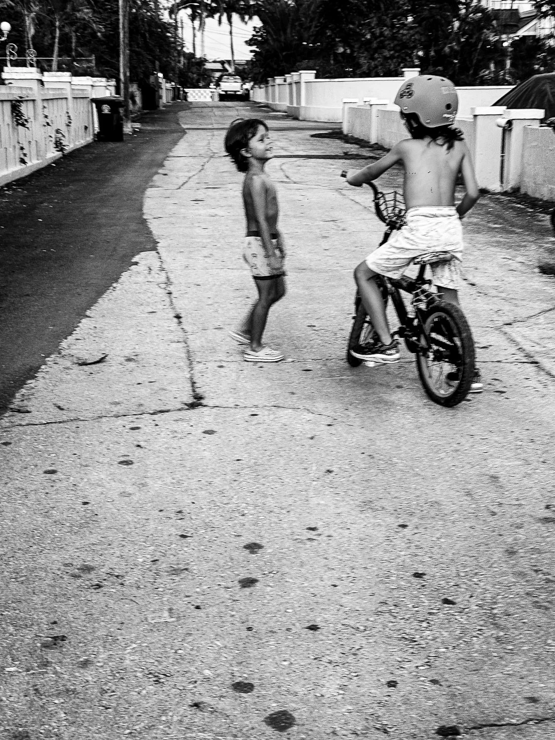 Two children, a boy on a bicycle with a helmet and a girl, stand on a cracked concrete street in a residential neighborhood, with fences, cars, and trees in the background.