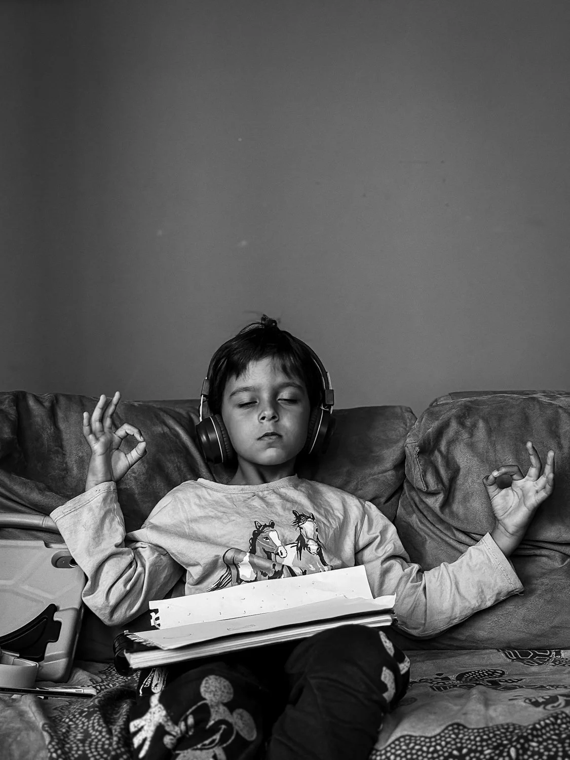 A young child with short hair wearing a graphic long-sleeve shirt and pajama pants, sitting on a couch with eyes closed, headphones on, and making an 'OK' hand gesture with both hands, holding a book on their lap.