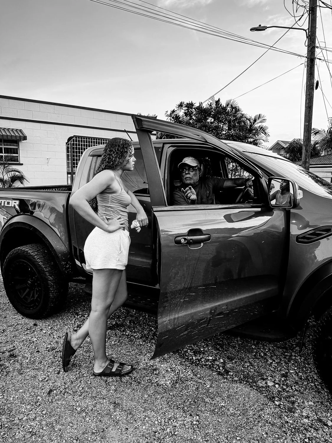 A woman with curly hair, wearing a tank top, shorts, and sandals, stands next to a pickup truck with an open door. A man with glasses, a cap, and a beard is sitting inside the truck, pointing or gesturing. A young girl is visible in the passenger sea