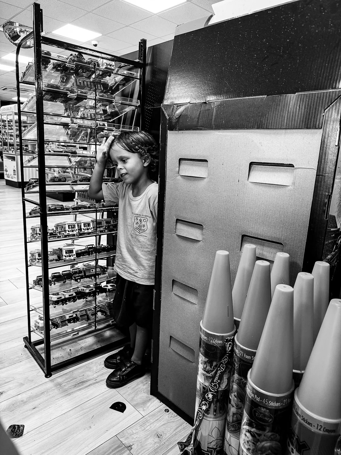 A young boy with a worried expression holding his head in a toy store aisle filled with toy cars and cones.