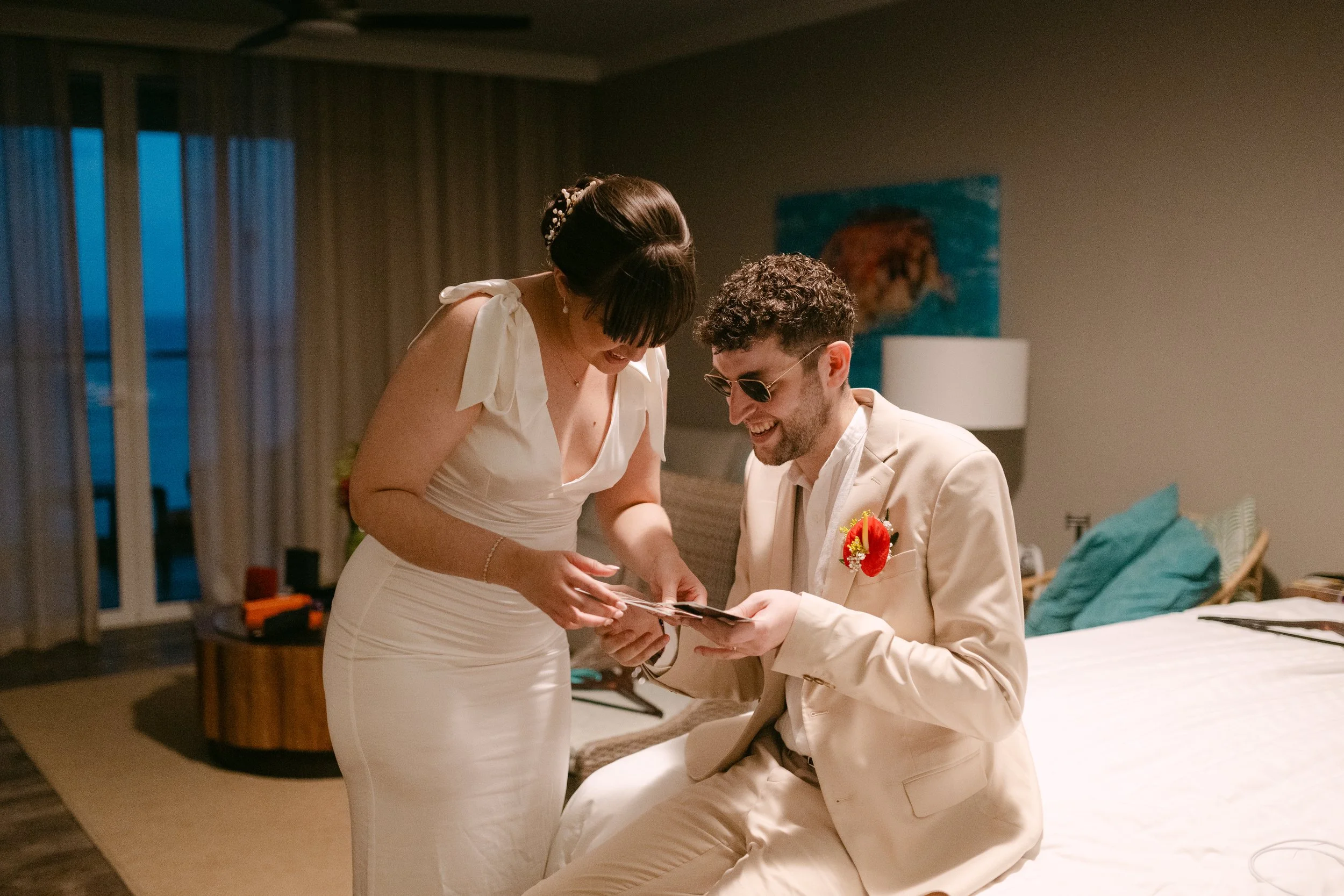 A man in a beige suit and sunglasses sitting on a bed, smiling, as a woman in a white dress shows him a photo or card. They are in a warmly lit room, with a painting, lamp, and pillows visible in the background.
