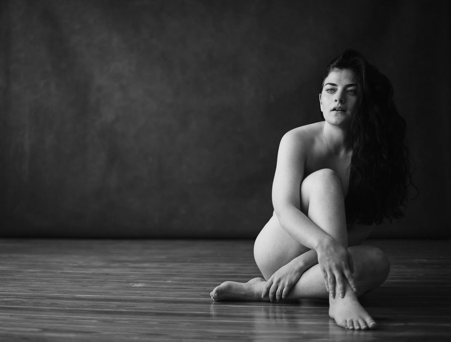 Black and white photo of a nude woman sitting on a wooden floor against a dark wall, with long wavy hair, looking contemplative.